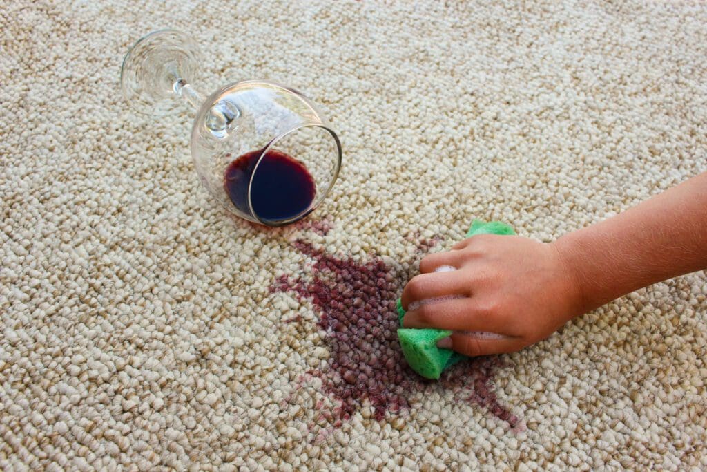 close shot of a glass of wine spilled on a carpet and a close shot of a hand scrubbing the stain with a sponge.