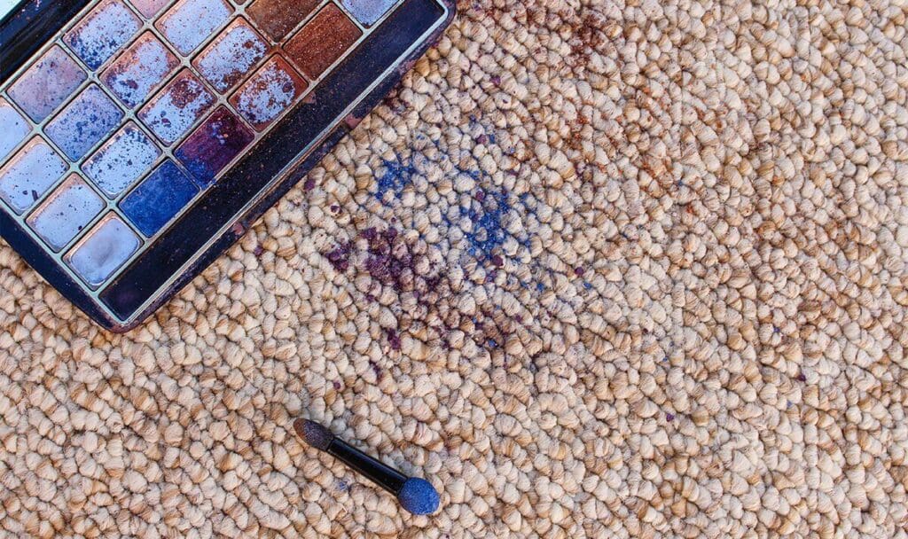 close shot of a makeup stain on carpet, makeup kit and a brush beside the stain.